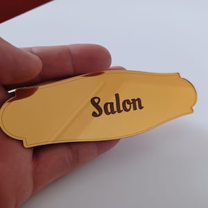 Pode incluir: Uma placa retangular dourada e reflexiva com bordas curvas decorativas. A palavra "Salon" está gravada em uma fonte escura, negrito e sem serifa no centro. A superfície é lisa e metálica, apresentando um reflexo nítido.