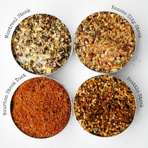 May include: Four small metal containers filled with different spice blends for steak. The blends are labeled "Montreal Steak", "Kansas City Steak", "Bourbon Steak Dust", and "Seattle Steak".