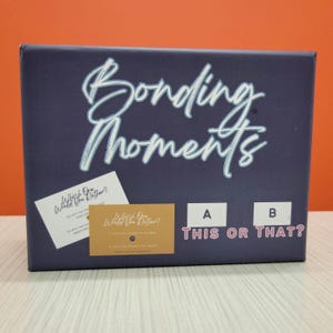 May include: A blue box with the words "Bonding Moments" in white script. The box has two white cards with the text "Which One Would You Rather?" and the words "This or That?" printed on the front.