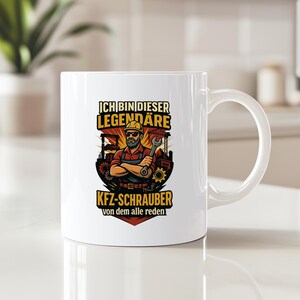 May include: White ceramic mug with a cartoon mechanic design, holding a wrench and wearing a hard hat. The text on the mug reads "ICH BIN DIESER LEGENDÄRE KFZ-SCHRAUBER von dem alle reden."