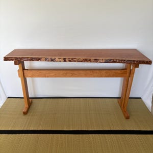 May include: A handcrafted wooden console table with a natural edge top. The table is made of light brown wood with a reddish-brown top. The legs are angled and supported by a horizontal beam. The table measures approximately 60 inches long.