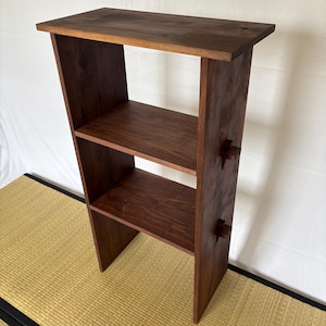May include: A dark brown wooden bookshelf with three shelves and a rectangular top. The bookshelf has a minimalist design with visible joinery details. The shelves are evenly spaced, and the wood grain is visible.