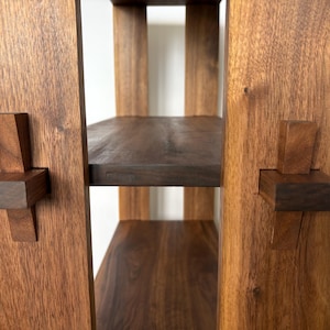 Peut inclure: Gros plan sur une &eacute;tag&egrave;re en bois avec trois tablettes. Les tablettes sont en bois fonc&eacute; et ont une finition naturelle. L'unit&eacute; est construite avec une technique de menuiserie unique, o&ugrave; les tablettes sont maintenues en place par des chevilles en bois qui sont ins&eacute;r&eacute;es dans des trous dans les c&ocirc;t&eacute;s de l'unit&eacute;.