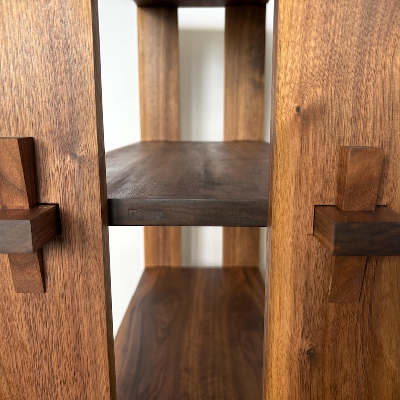 Peut inclure: Gros plan sur une &eacute;tag&egrave;re en bois avec trois tablettes. Les tablettes sont en bois fonc&eacute; et ont une finition naturelle. L'unit&eacute; est construite avec une technique de menuiserie unique, o&ugrave; les tablettes sont maintenues en place par des chevilles en bois qui sont ins&eacute;r&eacute;es dans des trous dans les c&ocirc;t&eacute;s de l'unit&eacute;.