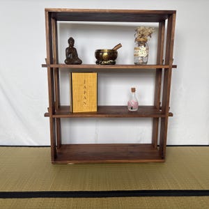 Peut inclure: Une &eacute;tag&egrave;re en bois brun fonc&eacute; &agrave; trois niveaux. Les &eacute;tag&egrave;res sont d&eacute;cor&eacute;es d'une petite statue de Bouddha en bronze, d'un bol chantant, d'un bocal en verre avec des fleurs s&eacute;ch&eacute;es, d'un livre intitul&eacute; "JAPAN" et d'une petite bouteille en verre avec un motif de fleur rose.