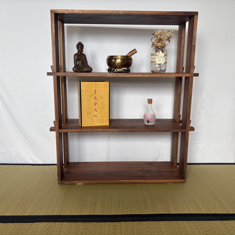 Peut inclure: Une &eacute;tag&egrave;re en bois brun fonc&eacute; &agrave; trois niveaux. Les &eacute;tag&egrave;res sont d&eacute;cor&eacute;es d'une petite statue de Bouddha en bronze, d'un bol chantant, d'un bocal en verre avec des fleurs s&eacute;ch&eacute;es, d'un livre intitul&eacute; "JAPAN" et d'une petite bouteille en verre avec un motif de fleur rose.