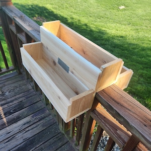 May include: A two-tiered wooden planter box, made of natural cedar wood, is mounted on a wooden deck railing. The planter box is designed to hold plants and flowers.