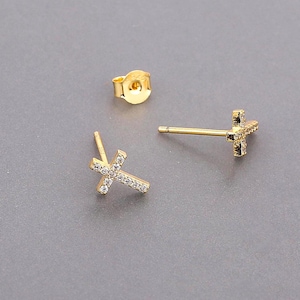 May include: A pair of gold-coloured stud earrings shaped like a cross, each with small, clear gemstones set into the cross. One earring back is also visible. The earrings are presented against a grey background.