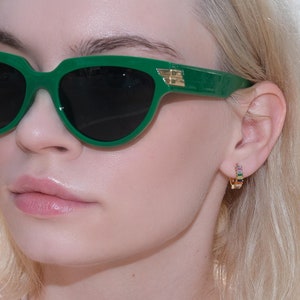 May include: Green cat-eye sunglasses with black lenses and gold detailing on the arms. The sunglasses are worn by a person with blonde hair and a white top. The background shows a body of water and boats.