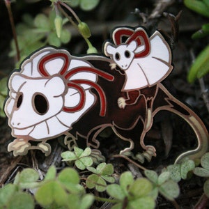 May include: A white and black enamel pin of two small animals wearing red and white bows. The larger animal is standing on all fours and the smaller animal is riding on its back.
