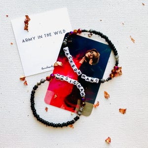 May include: Two black beaded bracelets with white letter beads spelling out "Jackson Wang" and "Magic Man". The bracelets are on a white surface with gold foil confetti.