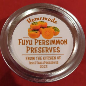 May include: A silver jar lid with a white label that reads "Homemade Fuyu Persimmon Preserves From the Kitchen of TreeTablePreserves 2023". The label features a cluster of orange persimmons.