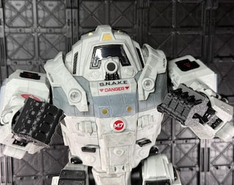 Peut inclure: Gros plan d'une figurine robotique blanche et grise avec des bras articulés noirs. La figurine porte l'inscription "S.N.A.K.E. DANGER" et le numéro "M7" sur son corps. Le robot est posé sur un fond industriel gris.