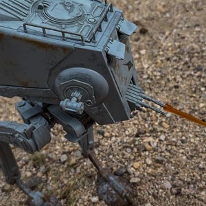 May include: A grey AT-ST model, a bipedal walker from a science fiction franchise, stands on a textured surface. The model has detailed features, including a top hatch, side guns, and a forward-facing cannon with an orange beam.
