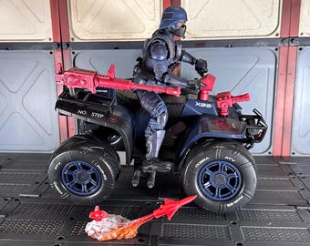 May include: A black and blue toy ATV with a red and black toy gun mounted on the front. A blue and black action figure is sitting on the ATV. The ATV has the words "COBRA" and "ATV" printed on the tires. The ATV also has the words "NO STEP" printed on the side. The ATV is on a black and gray tiled surface.