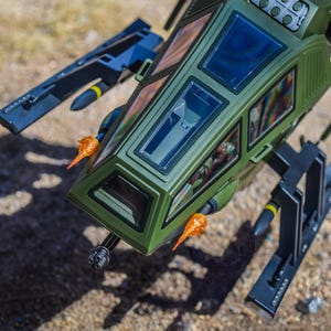 May include: A detailed overhead shot of a green toy helicopter with black and yellow missiles and orange flame effects. The helicopter has a cockpit with clear windows and a gun on the front. The toy is on a textured surface.