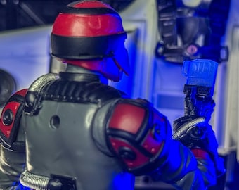 Peut inclure: Une figurine d'action futuriste avec un casque et des épaulettes rouges et noirs. La figurine est grise avec des accents rouges et un tube argenté reliant l'épaule à la tête. La figurine tient un objet bleu.