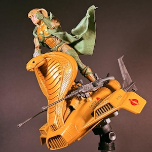 May include: A plastic action figure of a character wearing a green cape and cobra-themed headgear, standing on a yellow cobra-shaped vehicle. The vehicle features a long gun, a caution label reading CAUTION GUN REACTOR, and a red circular emblem.