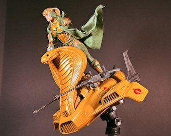 May include: A plastic action figure of a character wearing a green cape and cobra-themed headgear, standing on a yellow cobra-shaped vehicle. The vehicle features a long gun, a caution label reading CAUTION GUN REACTOR, and a red circular emblem.