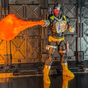 May include: Action figure of a futuristic warrior in a black and yellow suit, helmet with red visor, and a bright orange energy blast emanating from the right hand. The figure is posed against a metallic backdrop.