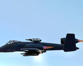 May include: A black military aircraft with a clear canopy and a gun turret on top. The plane has orange and purple accents on the tail and engines, and is set against a light blue sky.