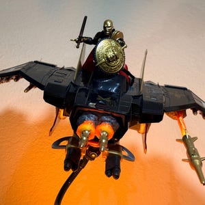 May include: A black and gold action figure riding a black winged aircraft. The figure holds a sword and a round gold shield. The aircraft features dual rear thrusters with orange flame effects, wing-mounted missiles, and gold accents.