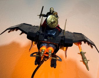 May include: A black and gold action figure riding a black winged aircraft. The figure holds a sword and a round gold shield. The aircraft features dual rear thrusters with orange flame effects, wing-mounted missiles, and gold accents.
