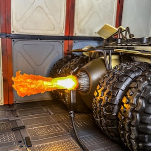 May include: Close-up of a black and gold vehicle with large, textured tires. A bright orange and yellow flame effect emanates from the vehicle's exhaust. The vehicle is connected to a black cable.