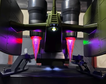 May include: A green toy spaceship with two glowing pink exhausts and a white light on the front. The spaceship is on a black surface.