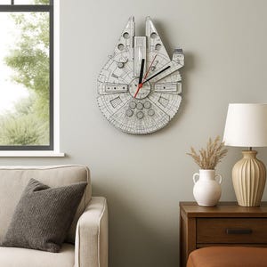 Puede incluir: Un reloj de pared con forma de la nave espacial Halcón Milenario. El reloj es de color gris claro con detalles en negro y manecillas rojas. Está montado en una pared gris claro.