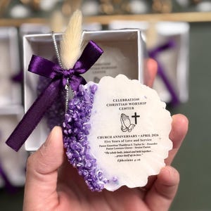 May include: A white, irregularly shaped ornament with purple accents and text, held in a small box. The ornament is decorated with purple crystals and a purple ribbon. The text reads "CELEBRATION CHRISTIAN WORSHIP CENTER" and "CHURCH ANNIVERSARY - APRIL 2026."