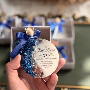 May include: A round, decorative ornament with blue and silver accents, a blue ribbon, and the text "Dail Louise 70th Celebrating a Life of Love & Grace". The ornament is held in a hand.