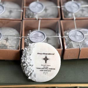 Puede incluir: Un recuerdo conmemorativo blanco de forma ovalada con el texto "Always Remembered" y una cruz. También se incluye el texto "Marquis Jaquez 1990-2025" y una cita. El recuerdo tiene un borde decorativo plateado y está empaquetado en una caja marrón.