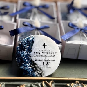 May include: A round, white and blue decorative item with text that reads "PASTORAL ANNIVERSARY Pastor Charles Lovett III & First Lady Chiquita Lovett 12th". The item has a cross symbol and is surrounded by gift boxes with blue ribbons.