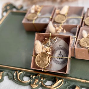 May include: Gift boxes with clear lids, each containing a round, white item with Arabic calligraphy and a gold wax seal featuring a mosque design. The boxes are decorated with dried flowers and gold accents, presented on a green tray.