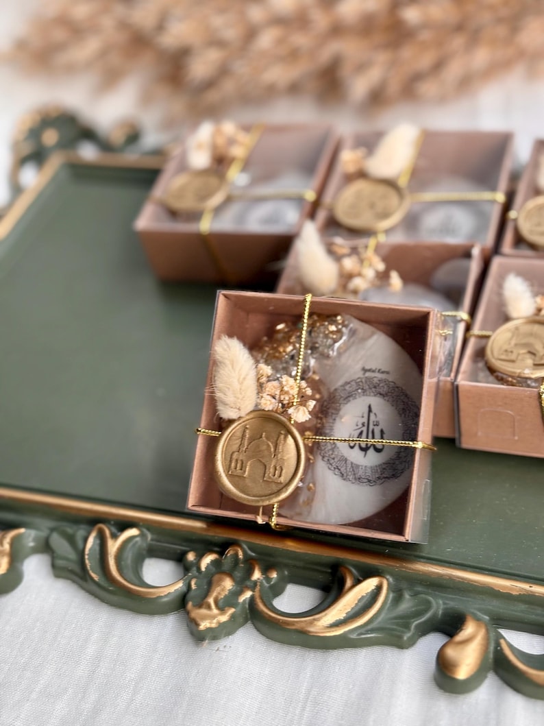 May include: Gift boxes with clear lids, each containing a round, white item with Arabic calligraphy and a gold wax seal featuring a mosque design. The boxes are decorated with dried flowers and gold accents, presented on a green tray.
