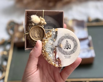 May include: A decorative, round resin coaster with gold leaf accents and Arabic calligraphy. The coaster is held in a hand, with a small gift box in the background. The box has a wax seal with Arabic text.