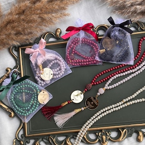 May include: An assortment of beaded necklaces and prayer beads in various colors, including red, white, green, and black. Some are packaged in sheer bags with ribbon ties. Gold-colored pendants and tassels are also visible.