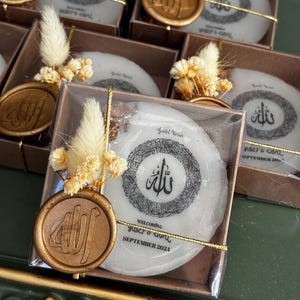May include: Wedding favour boxes, each containing a white, circular item with black calligraphy and a gold seal. The text "Welcoming Yuser & Gonul September 2024" is printed on one. Each box is decorated with dried flowers and gold string.