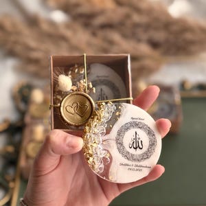 May include: A round, white and gold coaster with Arabic calligraphy and the text "Ayatul Kursi" and "Shakhboz & Shakhnobona 09.13.2025". The coaster is held in a small box with a gold seal.