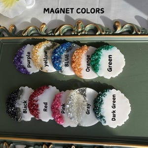 May include: A selection of decorative magnets in a range of colours: purple, yellow, blue, orange, green, black, red, pink, grey, and dark green. Each magnet has a white base and a textured, colourful accent. The text "MAGNET COLORS" is at the top.