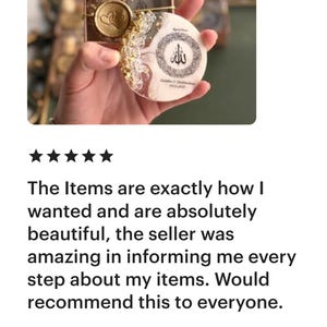 May include: A small, round decorative item with gold accents and a heart-shaped design, presented in a small box. The item has a white background with gold and glitter details. The item has Arabic text.