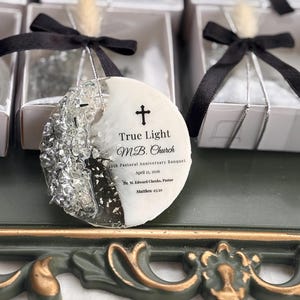 May include: A circular white and black resin ornament featuring silver glitter accents and a cross symbol. The text reads: True Light M.B. Church, 20th Pastoral Anniversary Banquet, April 25, 2026, Dr. M. Edward Cheeks, Pastor, Matthew 25:21.