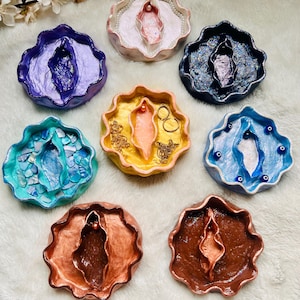 Puede incluir: Una colección de bandejas decorativas hechas a mano en varios colores, incluyendo azul, rosa y cobre. Cada bandeja presenta un diseño único con una forma central estilizada. Algunas bandejas tienen elementos decorativos como perlas o detalles dorados.