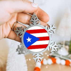 May include: A silver snowflake ornament displaying the Puerto Rican flag. The ornament has a circular centre with the flag's design of red and white stripes, a blue triangle, and a white star. The snowflake design has intricate cutouts.