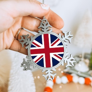 May include: A silver snowflake Christmas ornament featuring the Union Jack flag. The ornament is held up by a hand. The snowflake design has detailed cut-outs and is suspended from a silver chain.