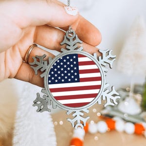 May include: A silver snowflake Christmas ornament featuring the American flag in the centre. The ornament is suspended by a silver chain. The background includes white and green festive decorations.