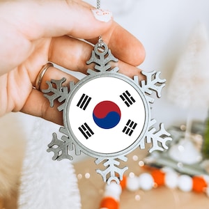 May include: A silver snowflake ornament displaying the South Korean flag. The ornament has a circular centre with the Taegeuk symbol and four trigrams. The snowflake design is detailed, suitable for festive decorations.