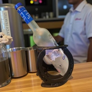 May include: A clear glass bottle of Grey Goose vodka with a blue label, held in a black horseshoe-shaped holder. The bottle is partially visible, with the label showing the brand name and logo. The holder sits on a wooden surface.