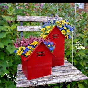 May include: Two red metal house-shaped planters filled with colorful flowers. The planters are 16.5cm/6.5in wide, 14.5cm/5.71in deep, and 20cm/7.87in tall. The flowers include yellow, blue, and purple blooms. The planters sit on a wooden surface.